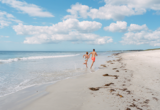Two people run along a sunny sandy beach by the sea with blue skies at Flower Camping Le Vorlen, Brittany.