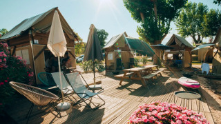 Safari-Zelt Airstay mit Holzterrasse, Blumenbeet und Gartenmöbeln unter Sonnenschirmen und Bäumen.
