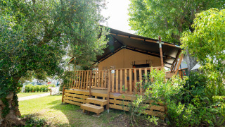 Safari-Zelt mit überdachter Holzterrasse, umgeben von grünen Bäumen und Pflanzen in der Sonne.