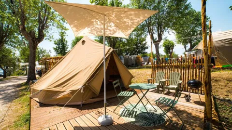 Safari Bell Tent med træterrasse, bord, stole, parasol og grill i naturskøn campingplads omgivet af træer.