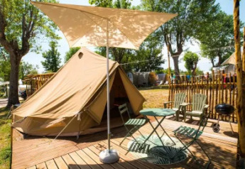 Tienda safari Bell Tent con terraza de madera, mesa, sillas, parasol y barbacoa en camping arbolado.
