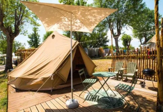 Tienda safari Bell Tent con terraza de madera, mesa, sillas, parasol y barbacoa en camping arbolado.
