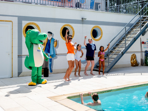 Niños y adultos bailan junto a la piscina con una mascota de cocodrilo en Flower Camping La Canadienne, Francia.