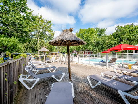 Sonnige Liegestühle und Strohdach-Sonnenschirme am Pool von Flower Camping La Canadienne, Nouvelle-Aquitaine.