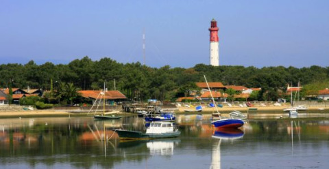 Scène côtière près d’Arès en Nouvelle-Aquitaine, France, avec des bateaux, des maisons et un phare à l’arrière-plan.