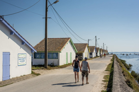 Zwei Menschen spazieren entlang einer Straße mit bunten Häusern am Wasser nahe Arès, Nouvelle-Aquitaine, Frankreich.