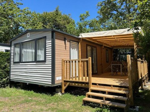 Stacaravan Premium 28m2 con terraza de madera cubierta, rodeada de árboles bajo un cielo soleado.