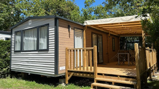 Stacaravan Premium 28m2 mobile home with covered wooden deck, surrounded by trees on a sunny day.