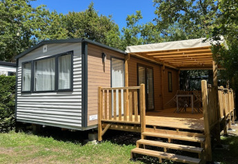Stacaravan Premium 28m2 mobile home with covered wooden deck, surrounded by trees on a sunny day.