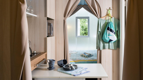 Moderne glampingaccommodatie met compacte keuken, eettafel en knusse slaapkamer gescheiden door gordijnen.