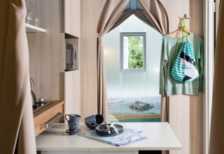 Moderne glampingaccommodatie met compacte keuken, eettafel en knusse slaapkamer gescheiden door gordijnen.