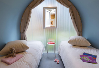 Chambre de glamping avec deux lits simples, peluche, livres pour enfants et petite fenêtre ouverte.