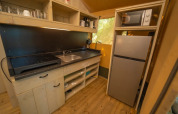 Angolo cucina glamping con piano cottura, lavello, microonde, frigorifero, scaffali e pavimento in legno.