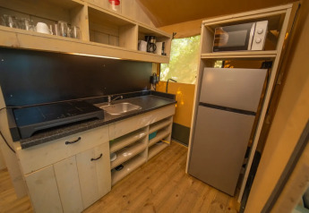 Glamping keuken met kookplaat, wasbak, magnetron, koelkast, open planken en houten vloer.