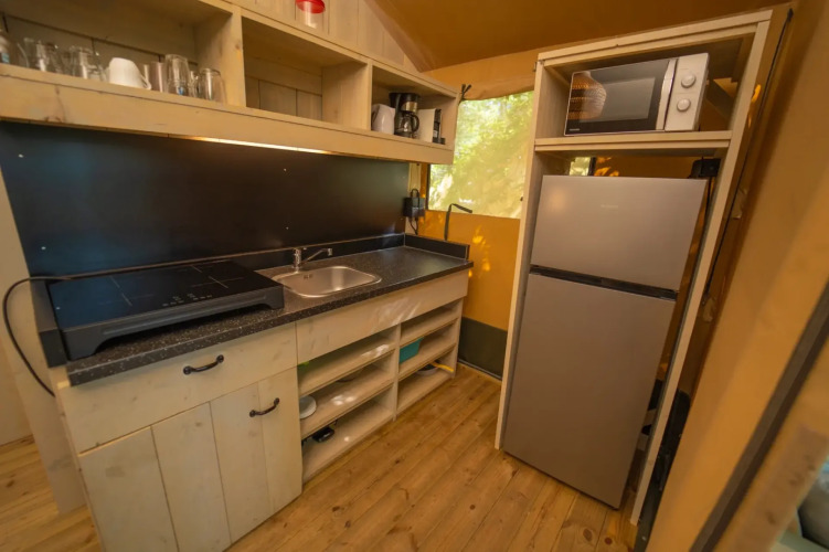 Acogedora cocina de glamping con estufa, fregadero, microondas, nevera, estantes y suelo de madera.