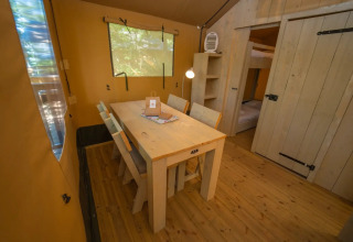 Intérieur d’une tente glamping avec table à manger en bois, quatre chaises et chambre visible au fond.