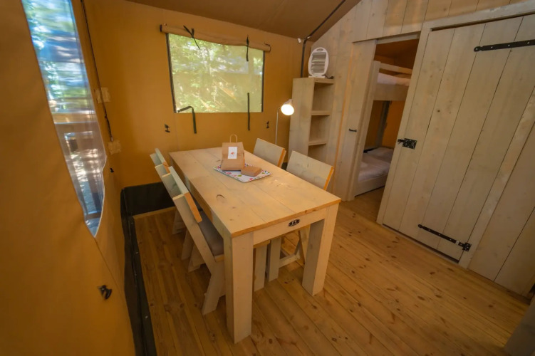 Interior de una tienda glamping con mesa de comedor de madera, cuatro sillas y dormitorio al fondo.