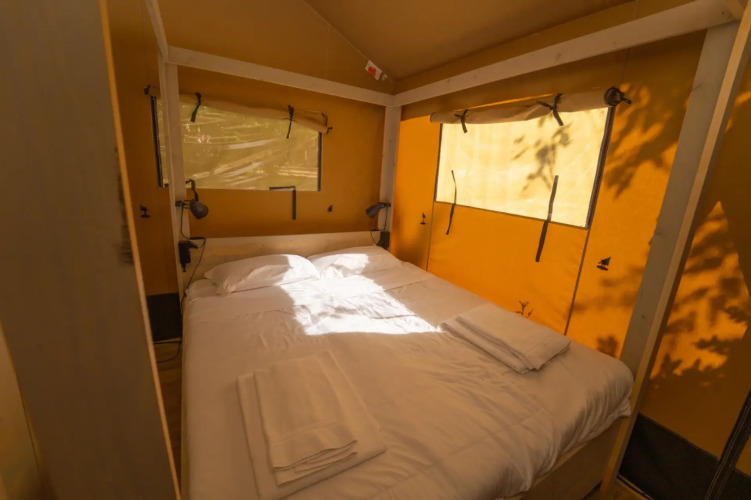 Interieur van een glamping-tent met tweepersoonsbed, witte lakens en zonlicht door ramen van canvas.
