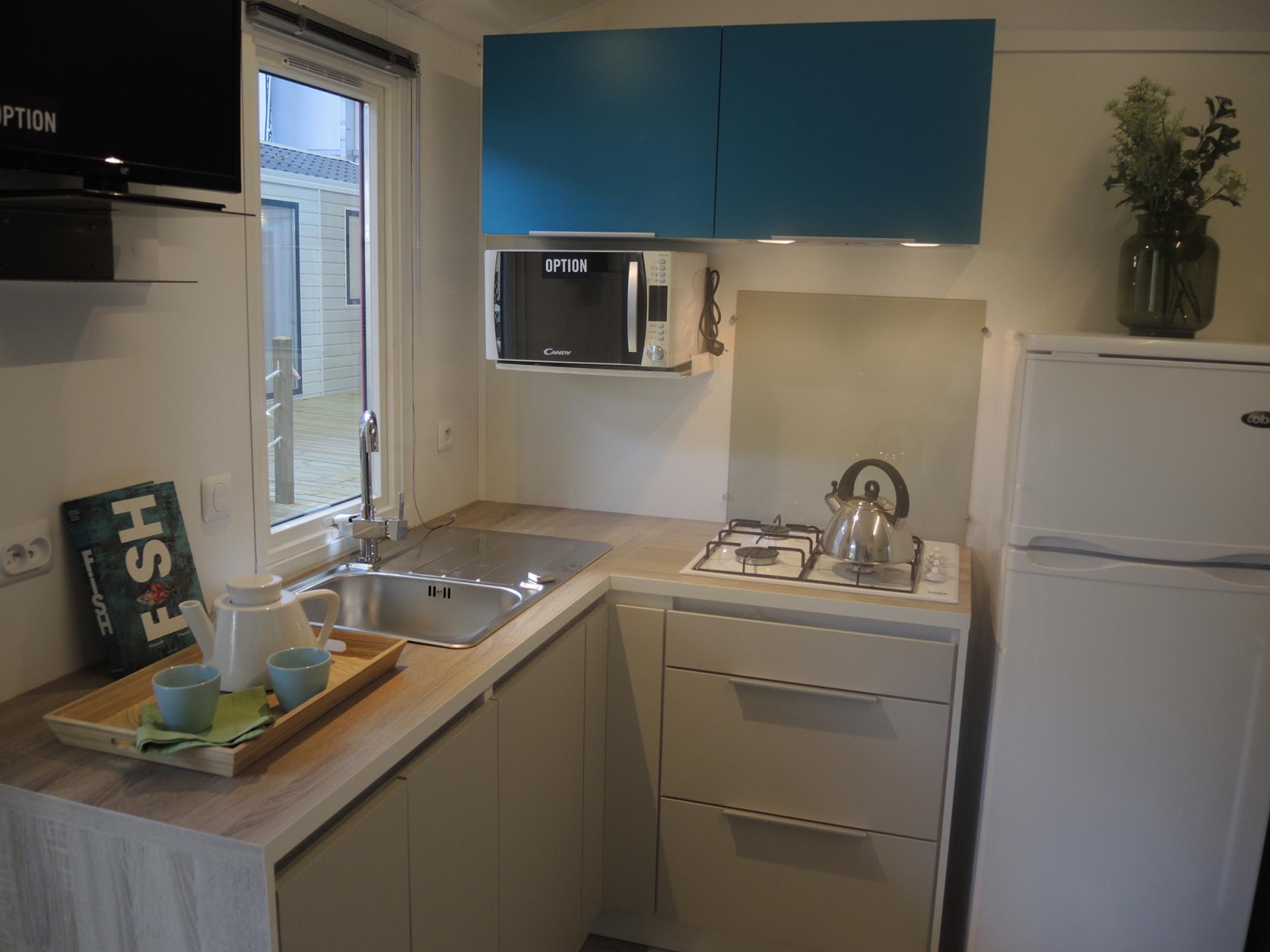 Moderne glampingkeuken met gootsteen, gaskookplaat, koelkast, magnetron, theeset en blauwe kasten.