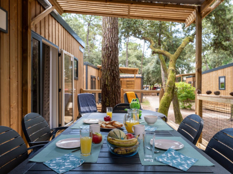 Outdoor breakfast table set on a wooden terrace at Flower Camping Monplaisir, surrounded by cabins and trees.
