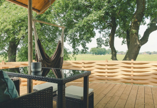 Terrasse en bois avec fauteuil hamac, table et chaises, vue sur champs verts et arbres aux Pays-Bas.