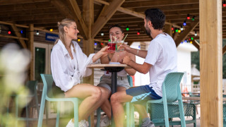 Tre venner nyder farverige drinks under overdækket terrasse med lyskæder på Flower Camping Monplaisir.