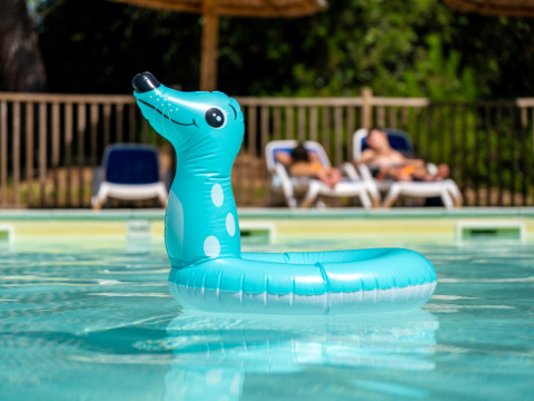Un flotador azul inflable en forma de animal flota en la piscina con tumbonas y gente al fondo.