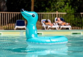 En blå, oppustelig badedyr flyder i en pool med solsenge og solbadende gæster i baggrunden, feriepark.