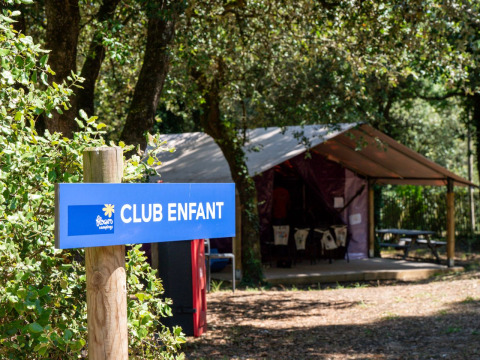 Blaues Schild mit 'CLUB ENFANT' vor einem Kinderclub im Wald bei Flower Camping Monplaisir, Frankreich.