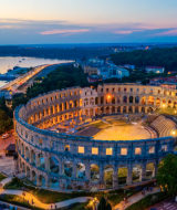 Amfiteatret i Pula nær Pomer, Istrien, Kroatien, ved solnedgang med bybelysning og udsigt til havnen.