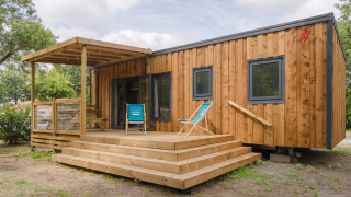 Photo of a modern wooden lodge with a porch, deck chairs, and steps set in a natural green environment.