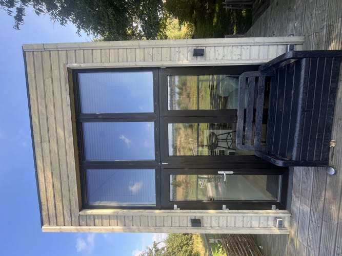 Casa pequeña moderna con grandes ventanales y terraza de madera, rodeada de naturaleza y cielo azul.