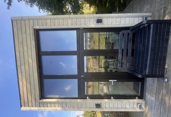 Casa pequeña moderna con grandes ventanales y terraza de madera, rodeada de naturaleza y cielo azul.