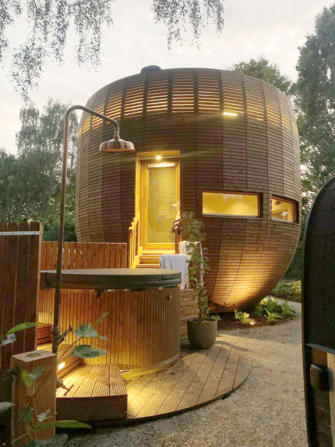 The round tiny house 'The Nutshell' made of wood, with a lit deck, hot tub, sauna, and outdoor shower at dusk.