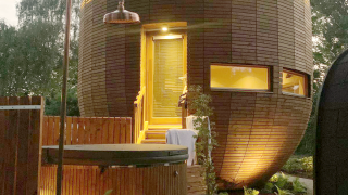La tiny house ronde en bois 'The Nutshell' avec terrasse éclairée, sauna, jacuzzi et douche extérieure au crépuscule.