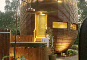 Das runde Minihaus 'The Nutshell' aus Holz mit beleuchtetem Whirlpool, Sauna und Außenbereich am Abend.