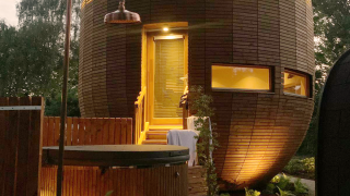 Ronde houten tiny house 'The Nutshell' met sfeerverlichting, hot tub, sauna en buitendouche bij schemer.