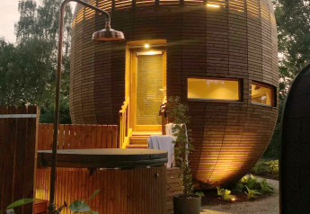 La tiny house redonda 'The Nutshell' de madera con jacuzzi iluminado, sauna y ducha exterior al anochecer.