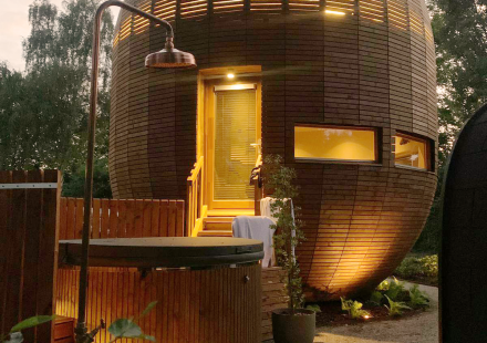 Rond houten tiny house 'The Nutshell' met verlichte patio, bubbelbad, sauna en buitendouche in de avond.