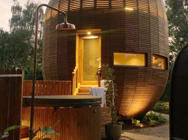 Rond houten tiny house 'The Nutshell' met verlichte patio, bubbelbad, sauna en buitendouche in de avond.