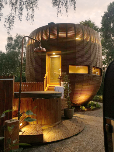La tiny house redonda 'The Nutshell' de madera con jacuzzi iluminado, sauna y ducha exterior al anochecer.