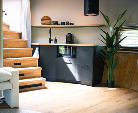 Kompakt køkkenhjørne med moderne sort inventar, trappe, plante og vindue i et tiny house ved Den Blanken.
