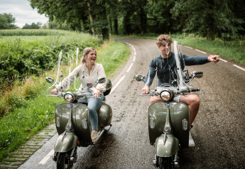 Zwei Menschen auf Motorrollern fahren lachend auf einer Landstraße bei The Nutshell in den Niederlanden.
