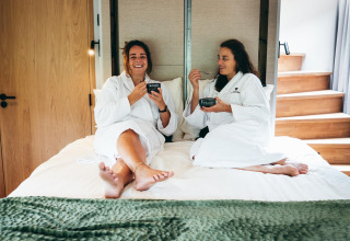 Twee vrouwen in badjassen genieten van ontbijt op bed in The Nutshell tiny house te Den Blanken, Nederland.