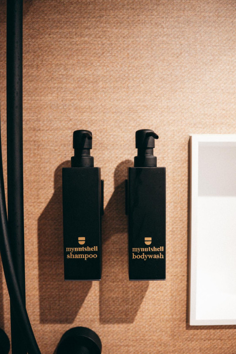 Twee zwarte flacons met opschrift 'mynutshell shampoo' en 'mynutshell bodywash' aan een badkamerwand.