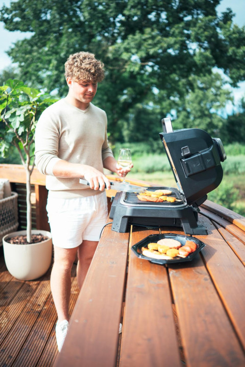 Een man barbequed op het terras van een tiny house met bubbelbad en sauna in Recreatiepark Den Blanken.