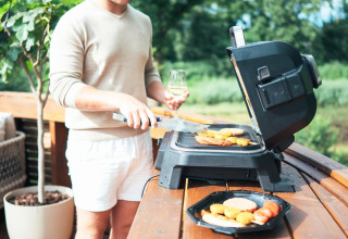 En mand griller mad på en terrasse ved et lille hus med boblebad og sauna i Recreation Park Den Blanken.