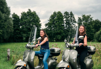 Zwei Frauen sitzen lächelnd auf Motorrollern vor grüner Landschaft beim Tiny House The Nutshell, Niederlande.