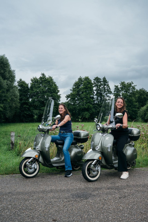 Twee vrouwen lachen op scooters voor groen landschap bij tiny house The Nutshell in Nederland.