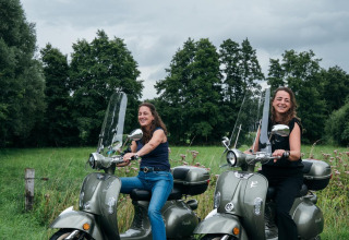 Twee vrouwen lachen op scooters voor groen landschap bij tiny house The Nutshell in Nederland.